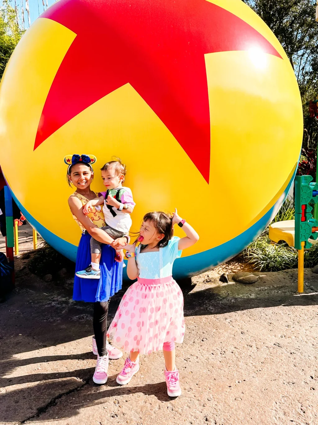 Three children pose happily in front of the giant Pixar Luxo Ball at Toy Story Land in Disney’s Hollywood Studios. The oldest girl wears a Jessie dress and holds her baby brother dressed as Buzz Lightyear, while the younger sister is dressed as Bo Peep in a pink polka-dot skirt and blue top strikes a playful pose. Bright sunlight, colorful outfits, and the iconic yellow-red-blue ball create a fun, joyful Disney family vacation moment.