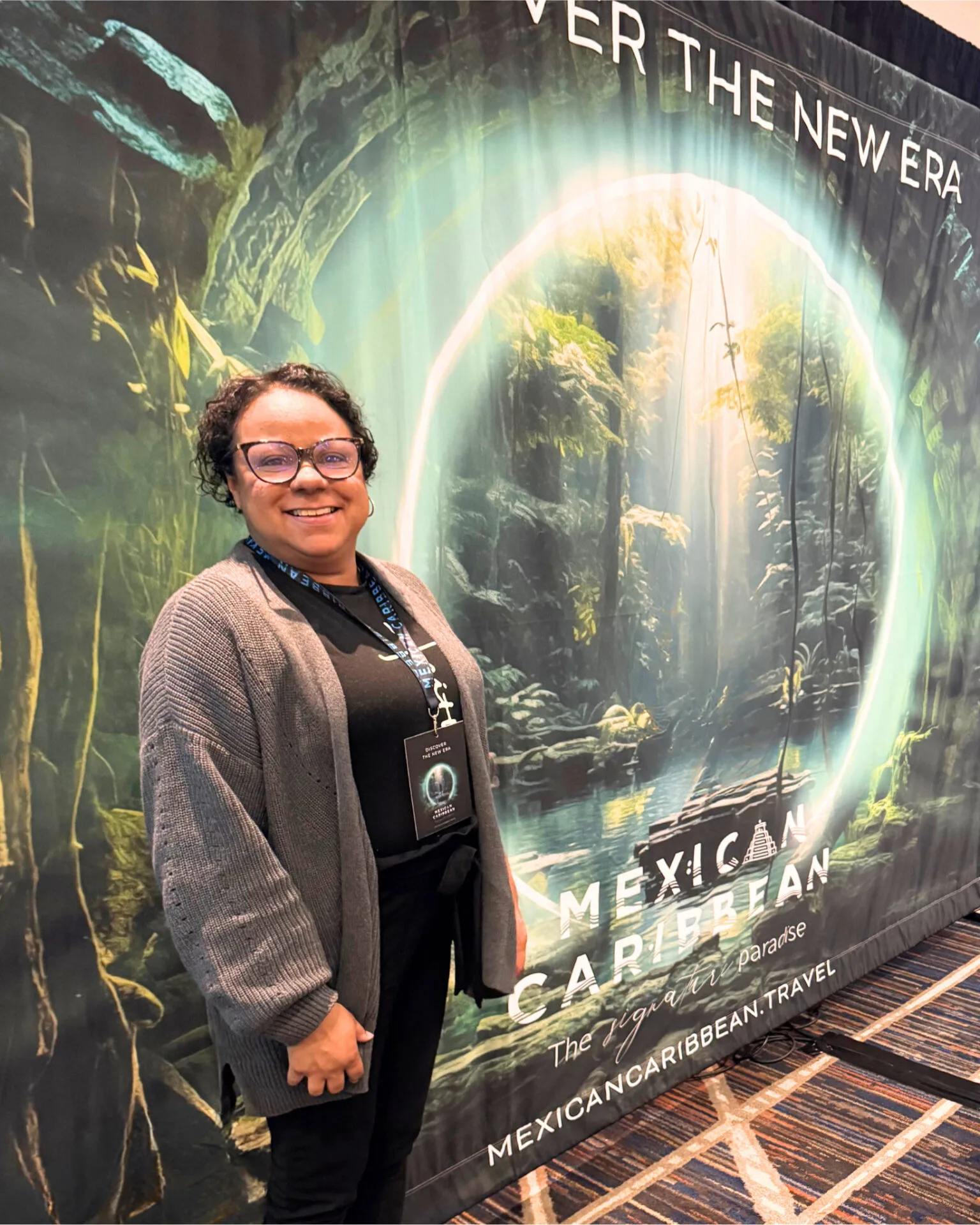 A smiling travel advisor (Joy) stands in front of a vibrant “Mexican Caribbean” event backdrop featuring a glowing cenote and lush jungle imagery. She wears glasses, a gray cardigan, and a conference badge while attending a Mexican Caribbean tourism event promoting travel to paradise destinations in Mexico